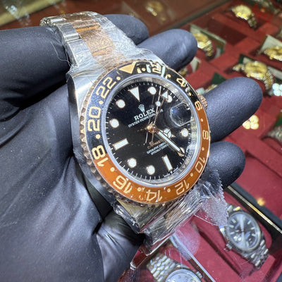 Luxury watch with black dial and orange bezel held by a gloved hand, with a display of other watches in the background.