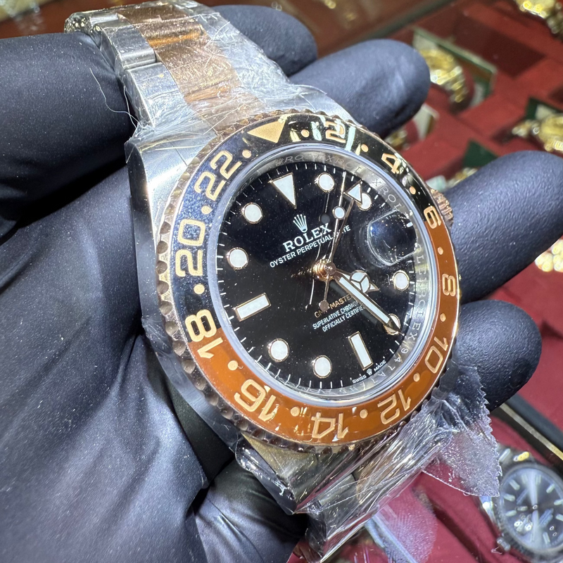 Luxury watch with black dial and orange bezel held by a gloved hand, with a display of other watches in the background.