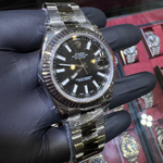 Gold and black watch held by a gloved hand with a display of watches in the background.