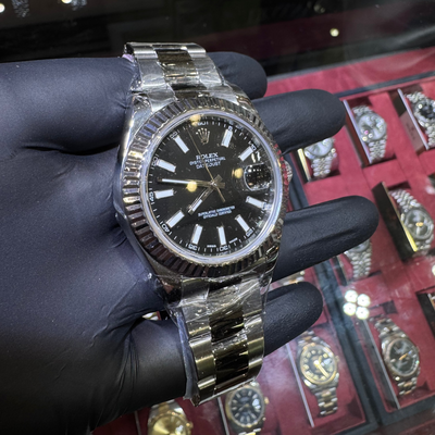 Gold and black watch held by a gloved hand with a display of watches in the background.