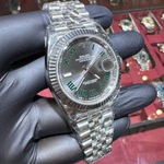 Silver watch with green Roman numeral dial held by a gloved hand against a jewelry display background.