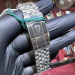 Silver watch with a Rolex logo on a wrist wearing a black glove, with a blurred background of jewelry.