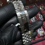 Close-up of a silver watch with a black strap held by a gloved hand, with a display of watches in the background.