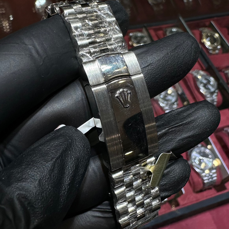 Close-up of a silver watch with a black strap held by a gloved hand, with a display of watches in the background.