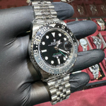 Silver watch with black dial held by a gloved hand against a display of watches.