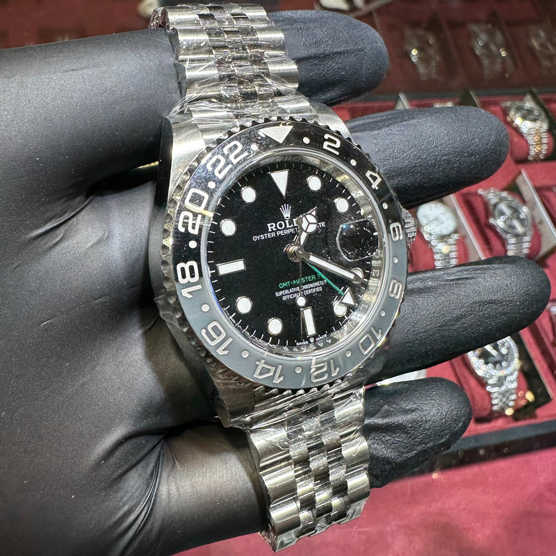 Silver watch with black dial held by a gloved hand against a display of watches.