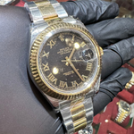 Luxury watch with black face and gold accents held by a gloved hand against a blurred jewelry store background.